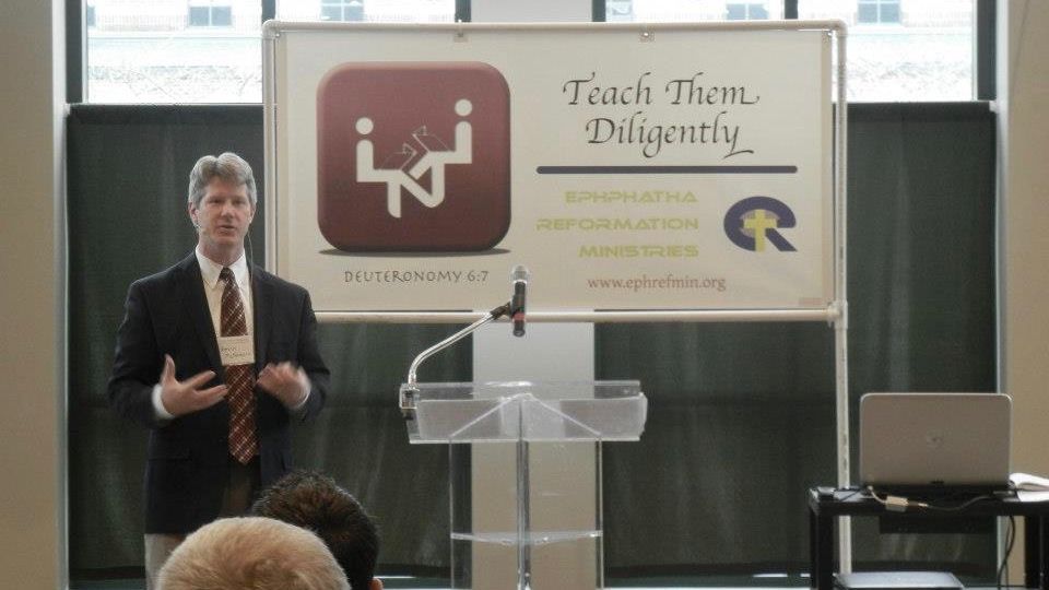 A speaker at a lectern in front of Teach Them Diligently and Ephphatha Reformation Ministries banners