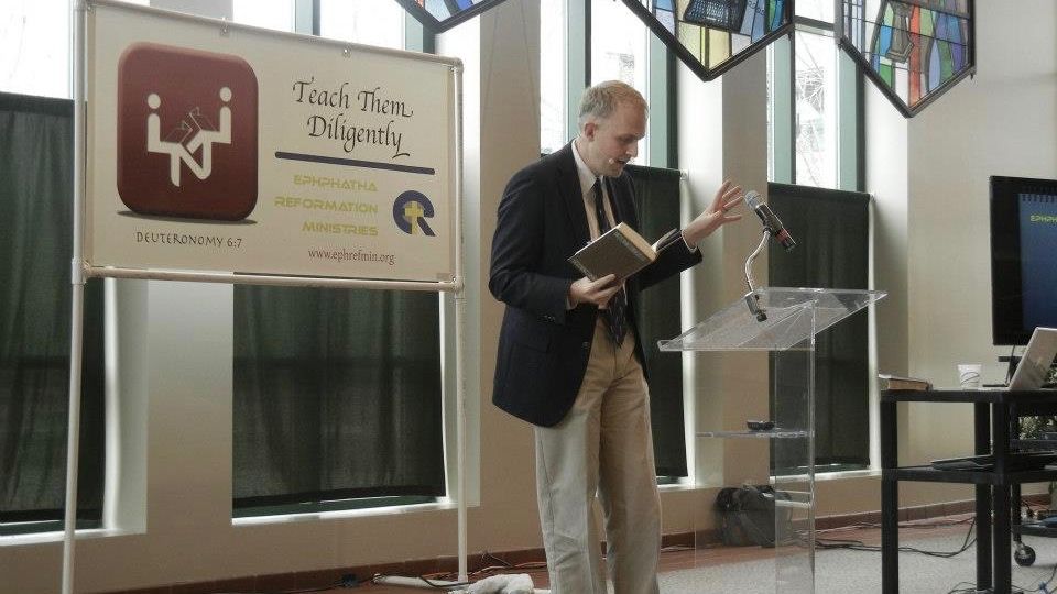 A speaker reads from a lectern beside a Teach Them Diligently conference banner
